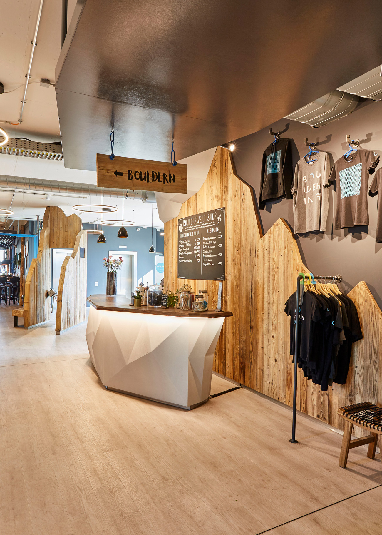 Foto: Empfangs- und Shopbereich mit geometrisch gestaltetem, weißen Tresen, Altholzoberflächen, integrierten Produktpräsentationen und moderner Beleuchtung in industrieller Architektur.
