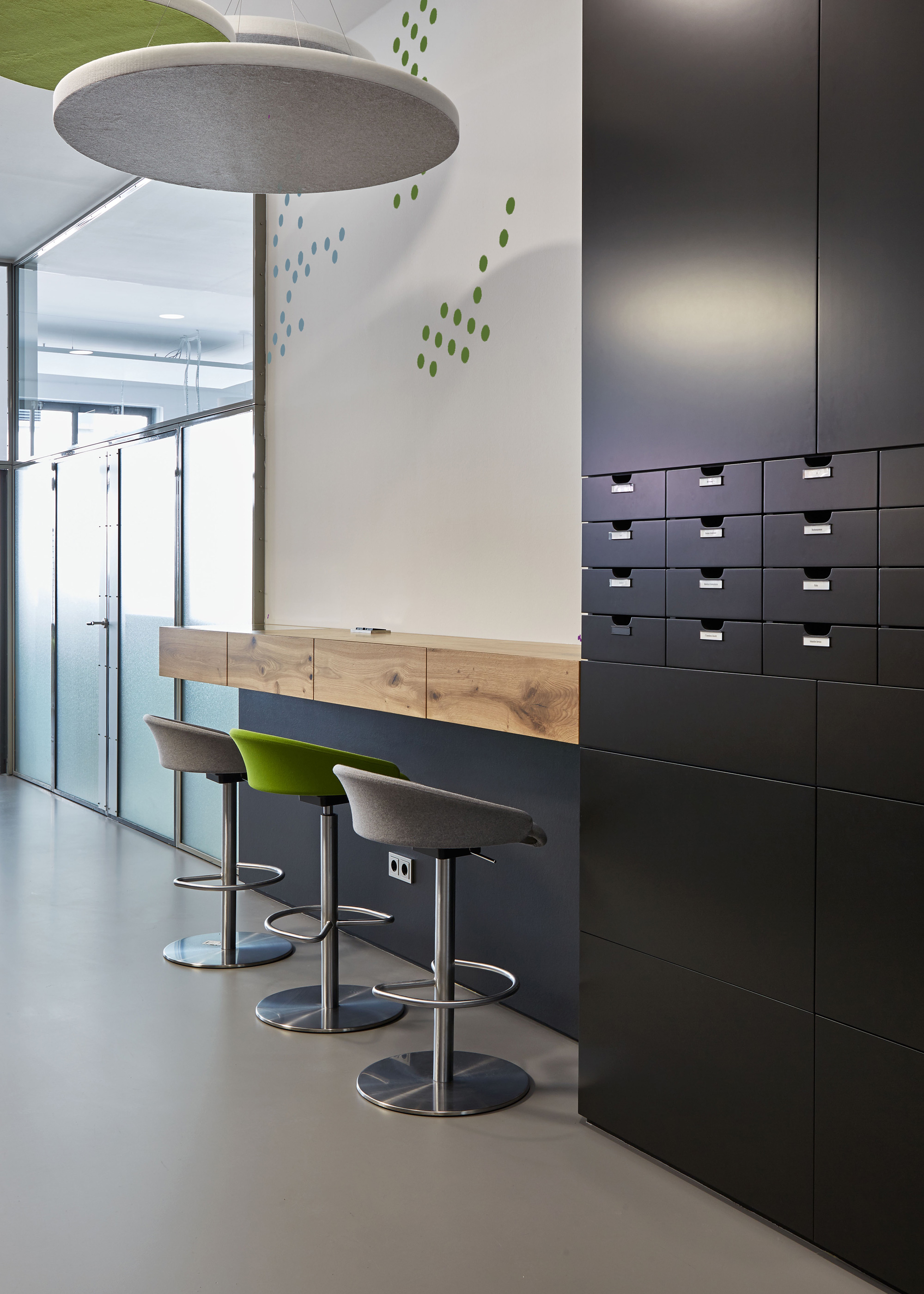 Foto: Moderner Aufenthaltsbereich mit Stehtisch, Barhockern in grün, grau und schwarzem Einbauschrank. Runden Akustikdeckenelementen und Wandfolierung in Punktmuster.