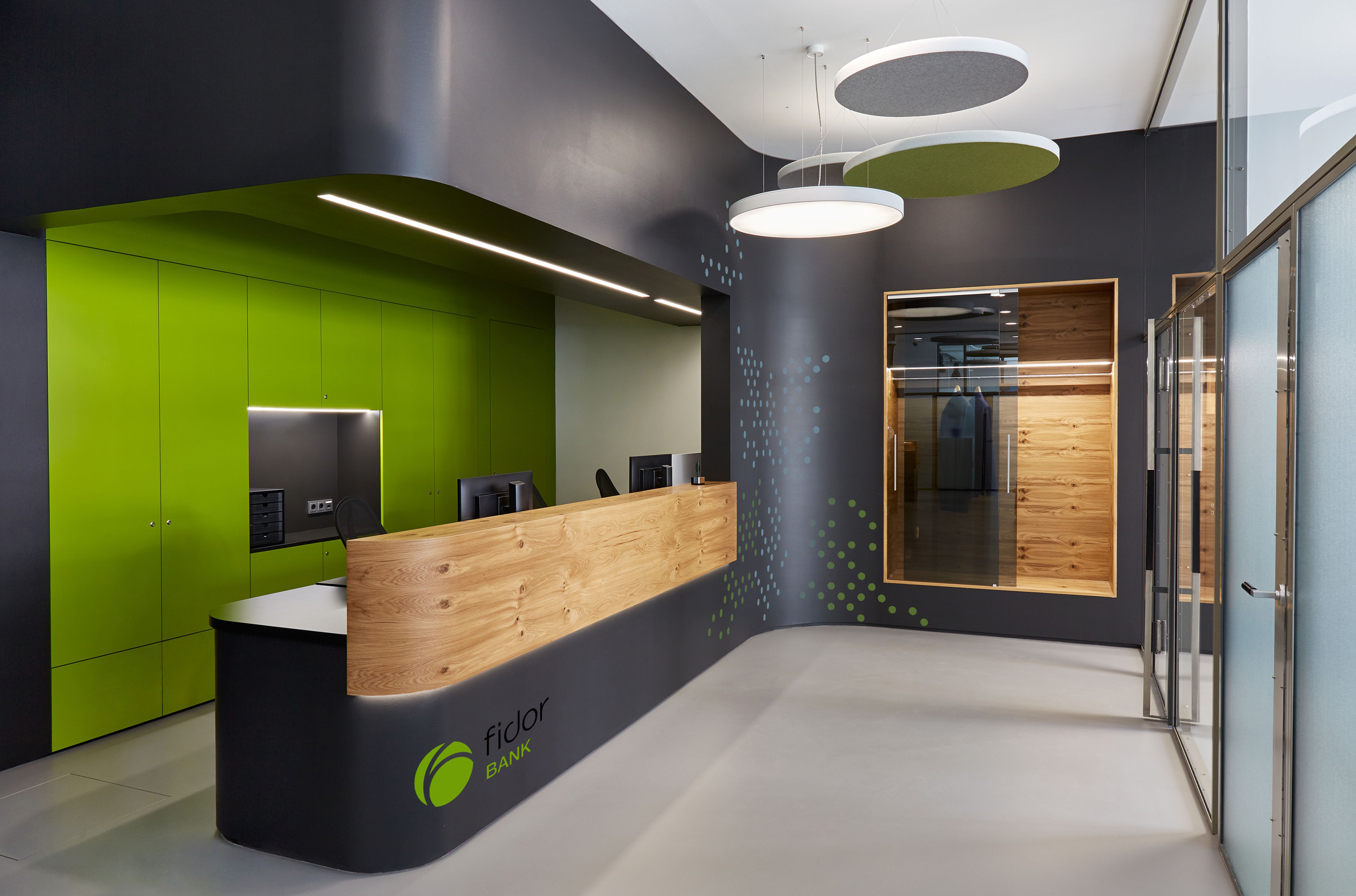 Foto: Empfangsbereich mit geschwungenen, schwarzen-grünen und holzverkleideten Einbauten, integrierten Regalen, Folierung in Punktmuster, grüne Akzente und runde Akustikelemente.