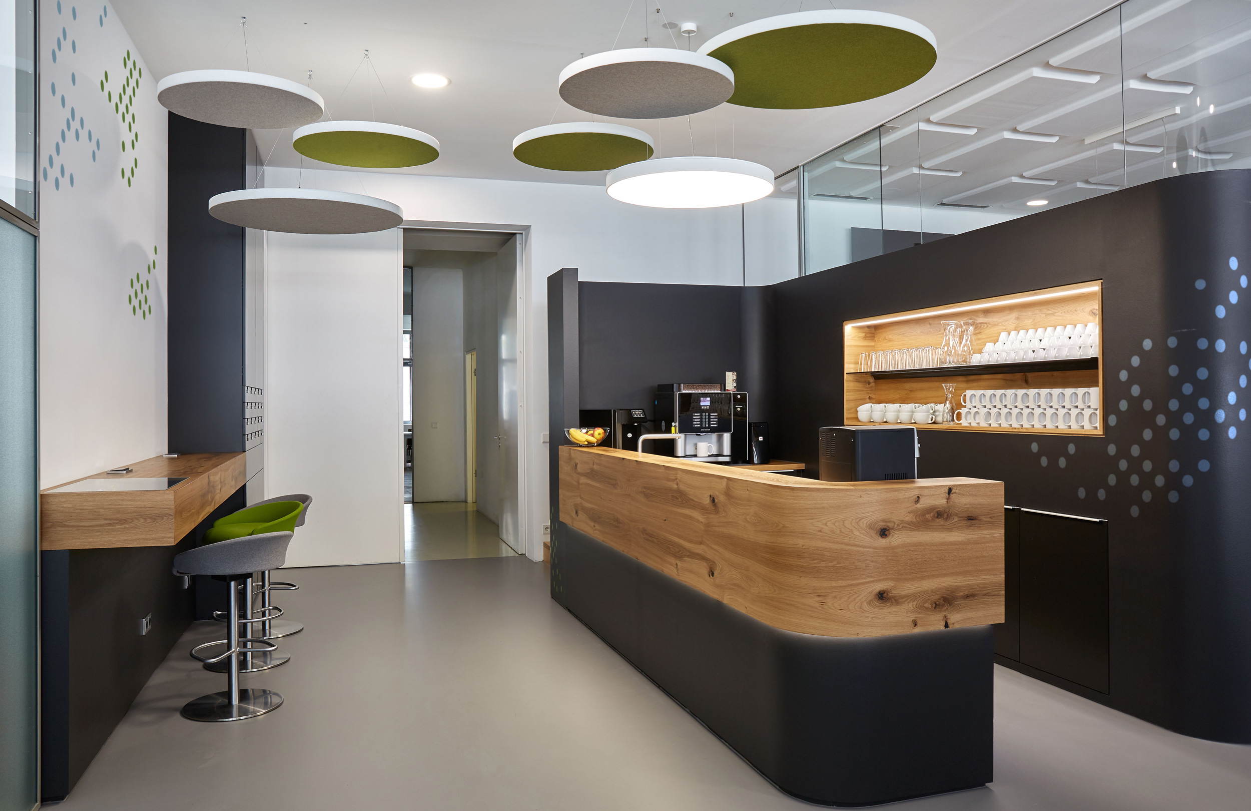 Foto: Modern gestaltete Baristabar mit  schwarzer, geschwungener Theke mit Eichenholzelementen, gegenüberliegender Stehtisch und runde weiß-grüne Deckenakustikelemente. 