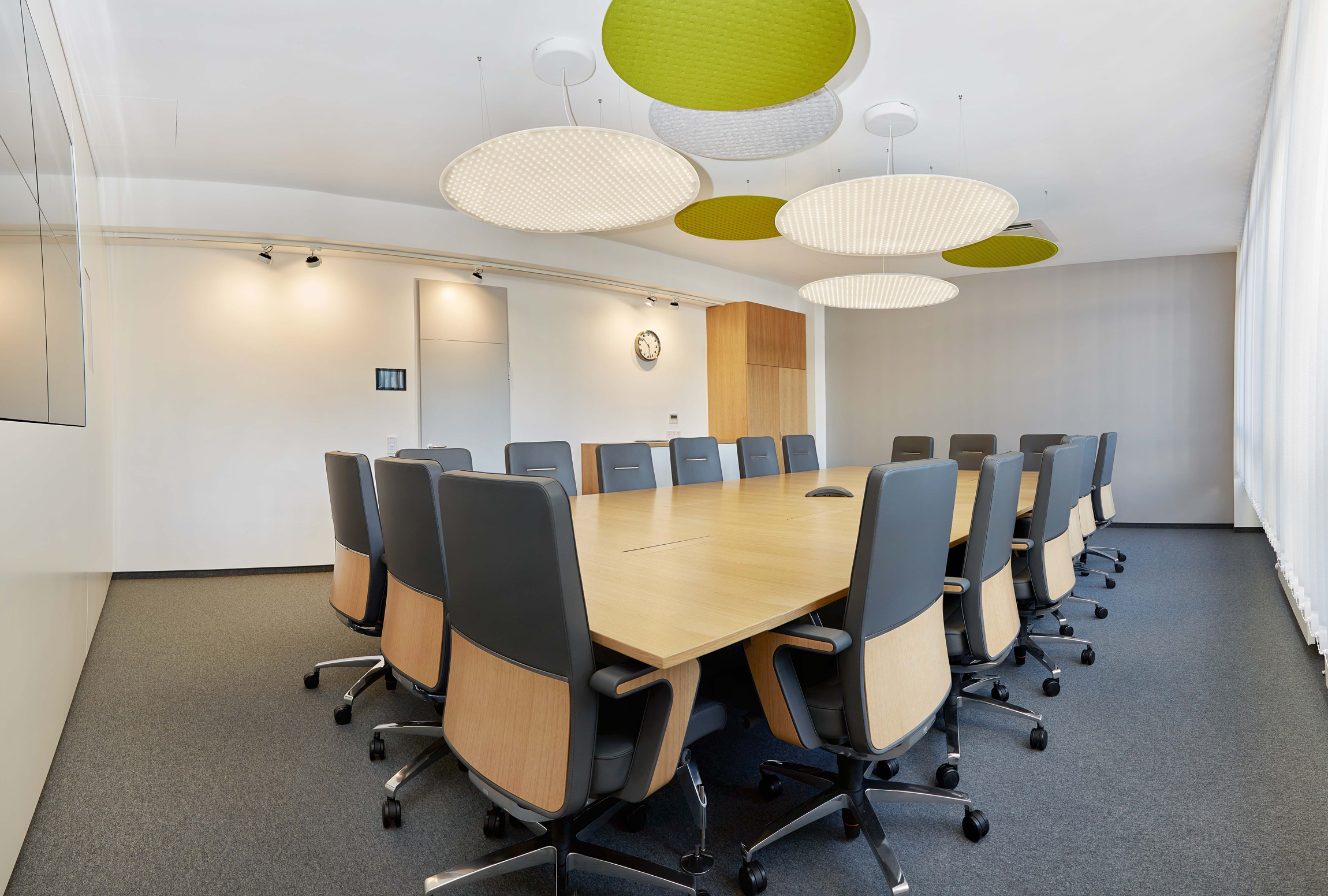 Foto: Moderner, heller Konferenzraum mit großem Holztisch, schwarzen Besprechungsstühlen, großem Bildschirm und runden grün-weißen Akustikelementen an der Decke.
