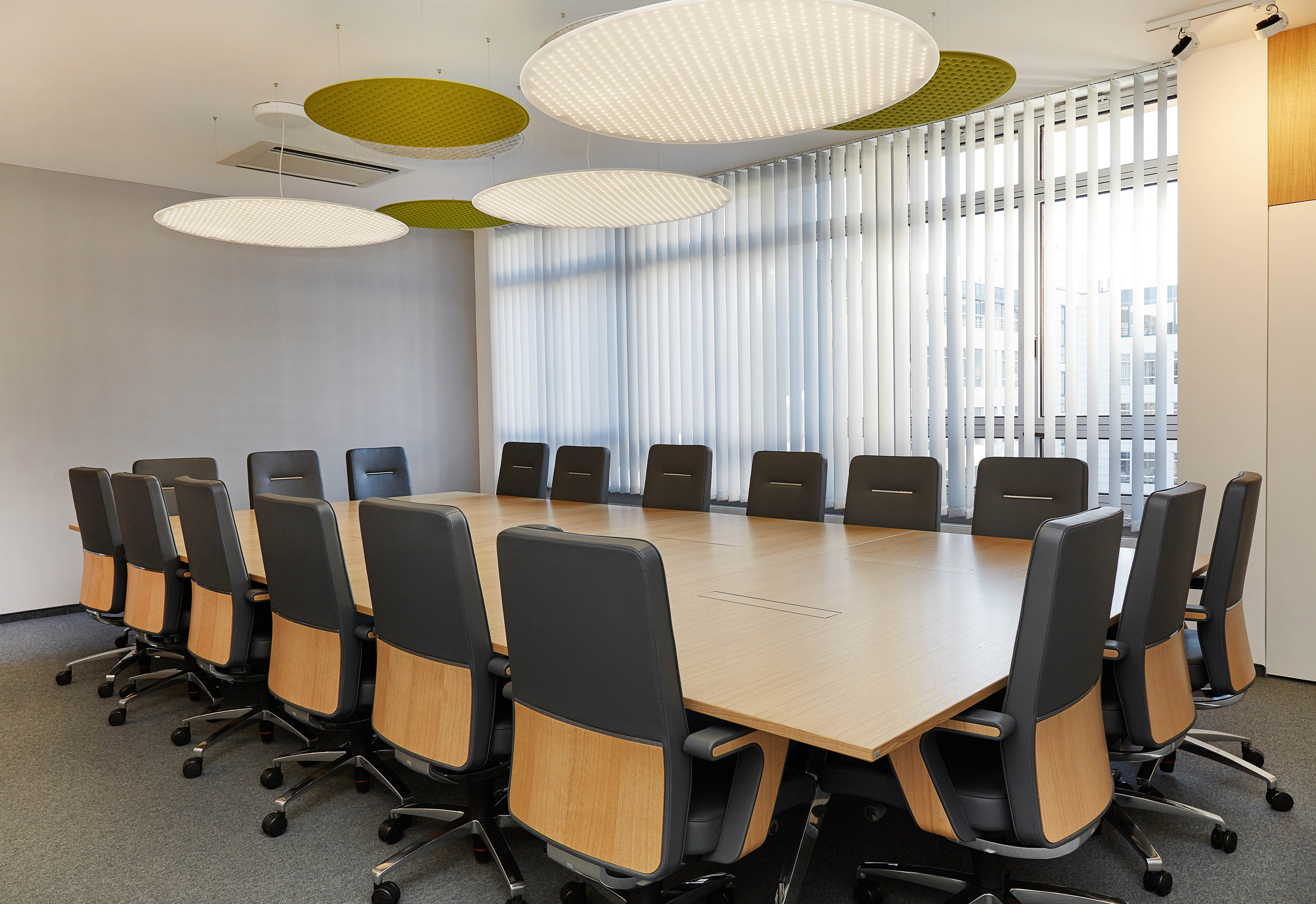 Foto: Moderner, heller Konferenzraum mit großem Holztisch, schwarzen Besprechungsstühlen, großem Bildschirm und runden grün-weißen Akustikelementen an der Decke.