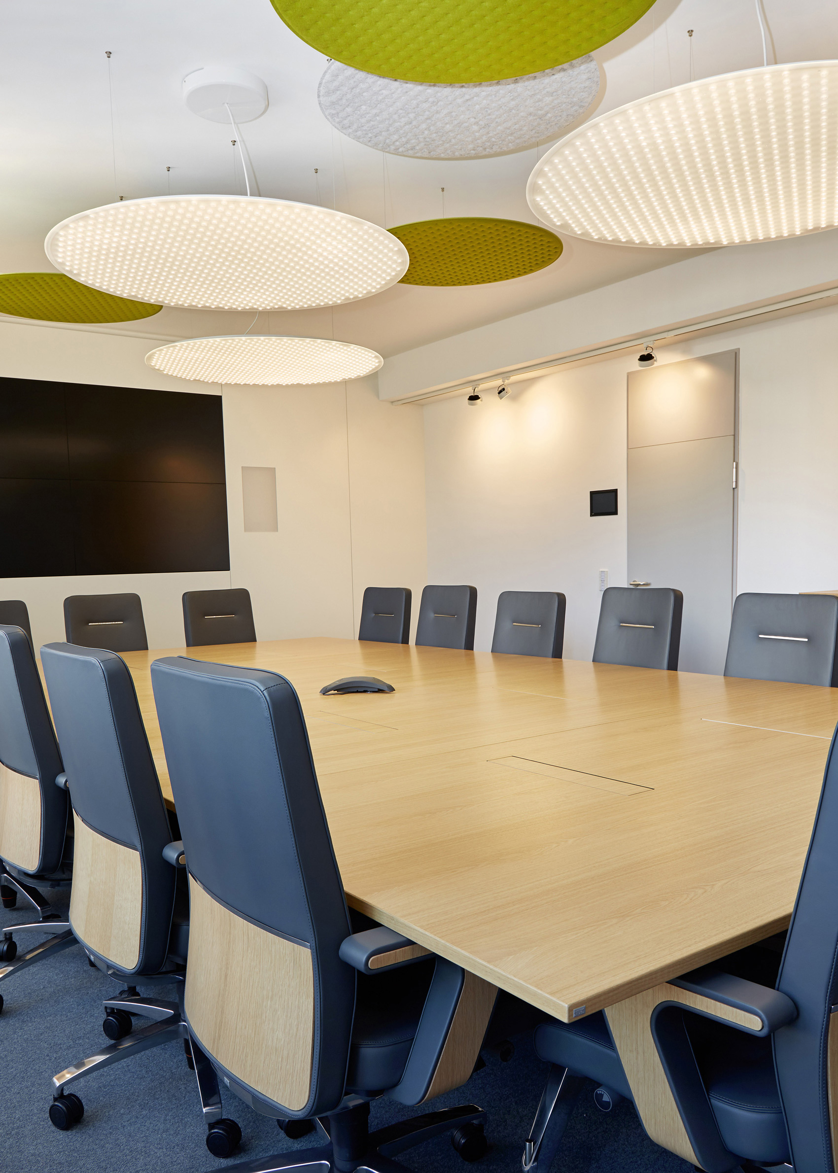 Foto: Moderner, heller Konferenzraum mit großem Holztisch, schwarzen Besprechungsstühlen, großem Bildschirm und runden grün-weißen Akustikelementen an der Decke.
