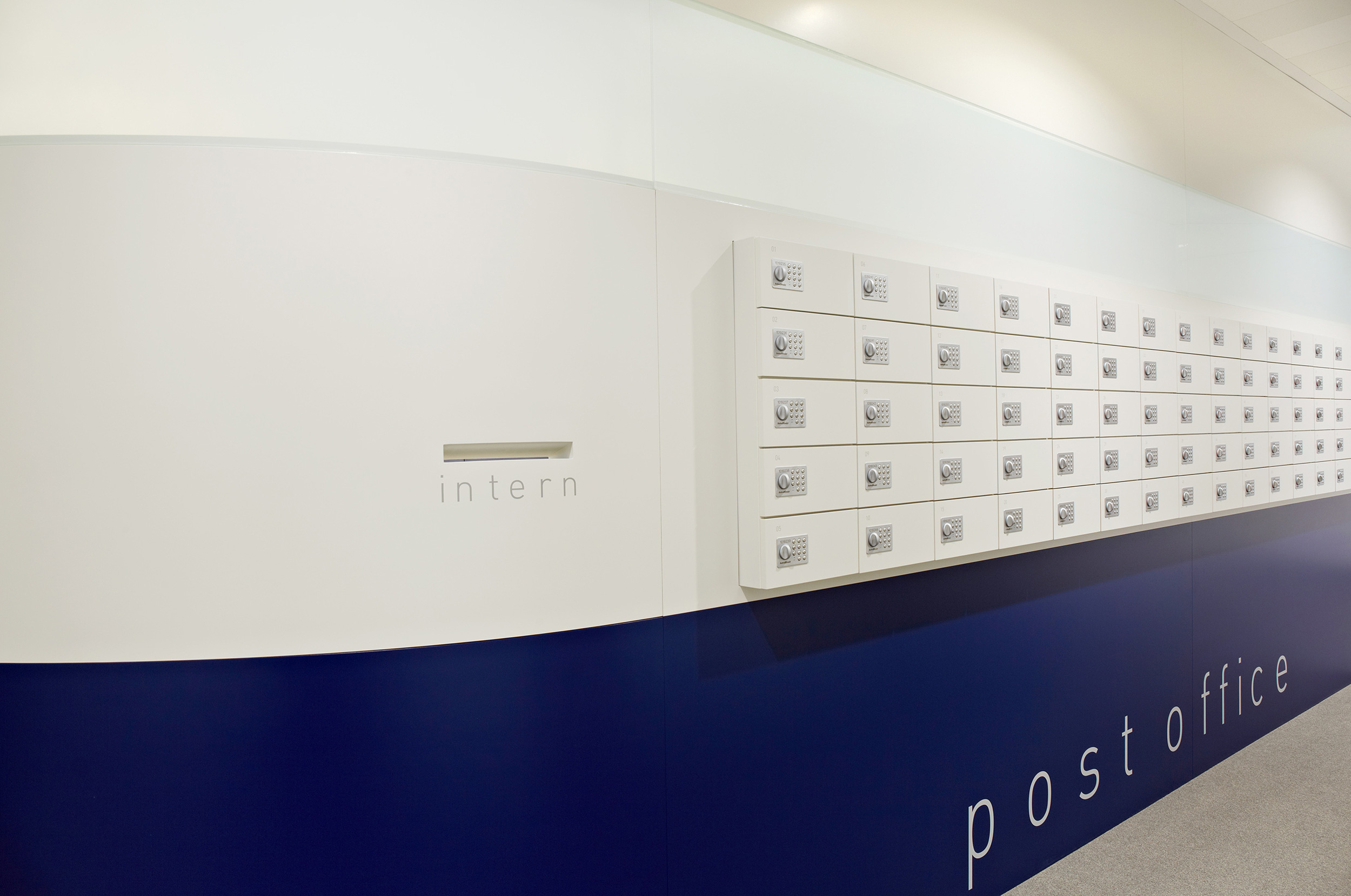Foto: Moderne Post- und Paketstation in einem Bürogebäude mit weißen Schließfächern an einer Seiten eines zentralen Raumelements, mit unterem Bereich in dunkelblau.