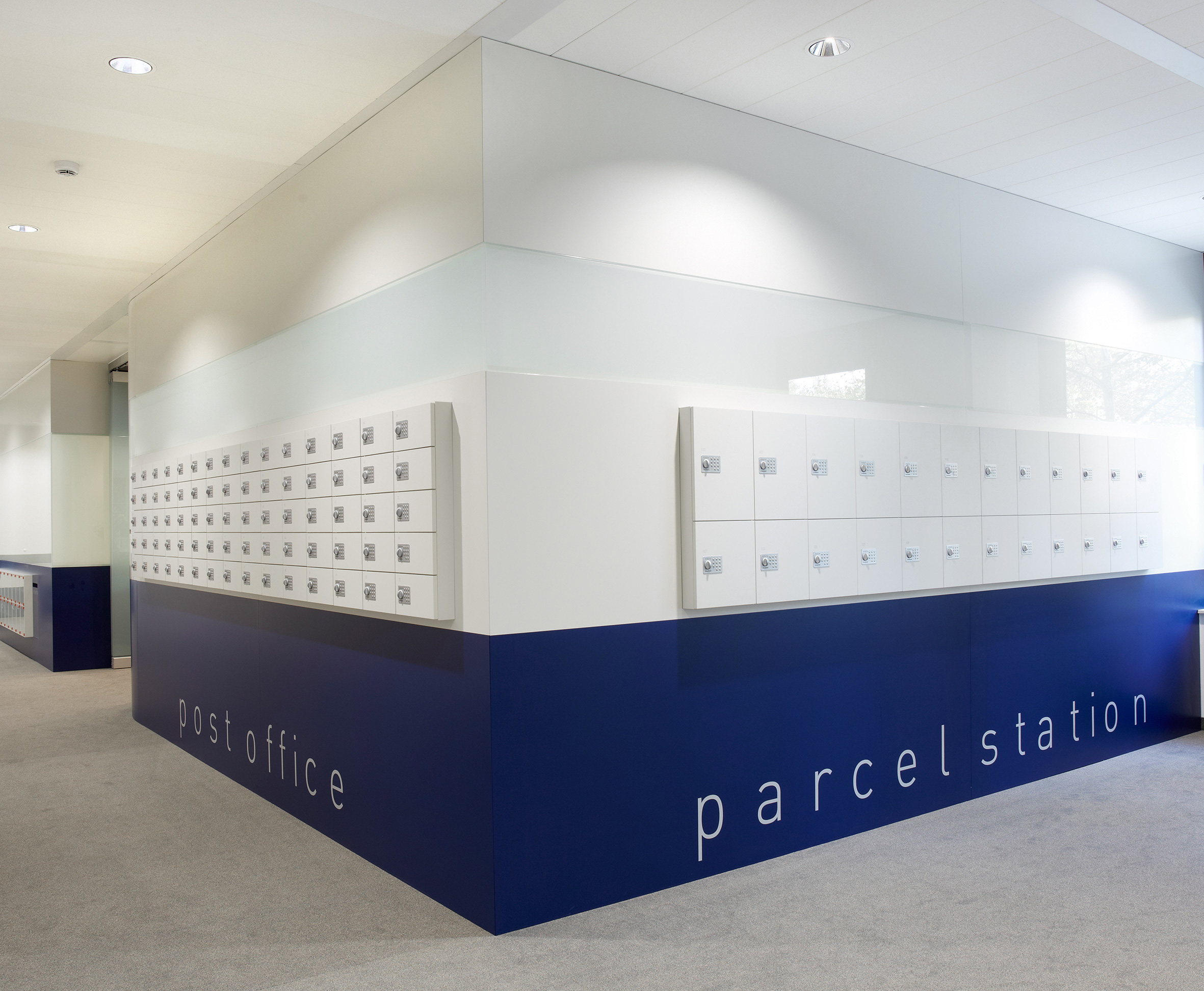 Foto: Moderne Post- und Paketstation in einem Bürogebäude mit weißen Schließfächern an zwei Seiten eines zentralen Raumelements, mit unterem Bereich in dunkelblau.