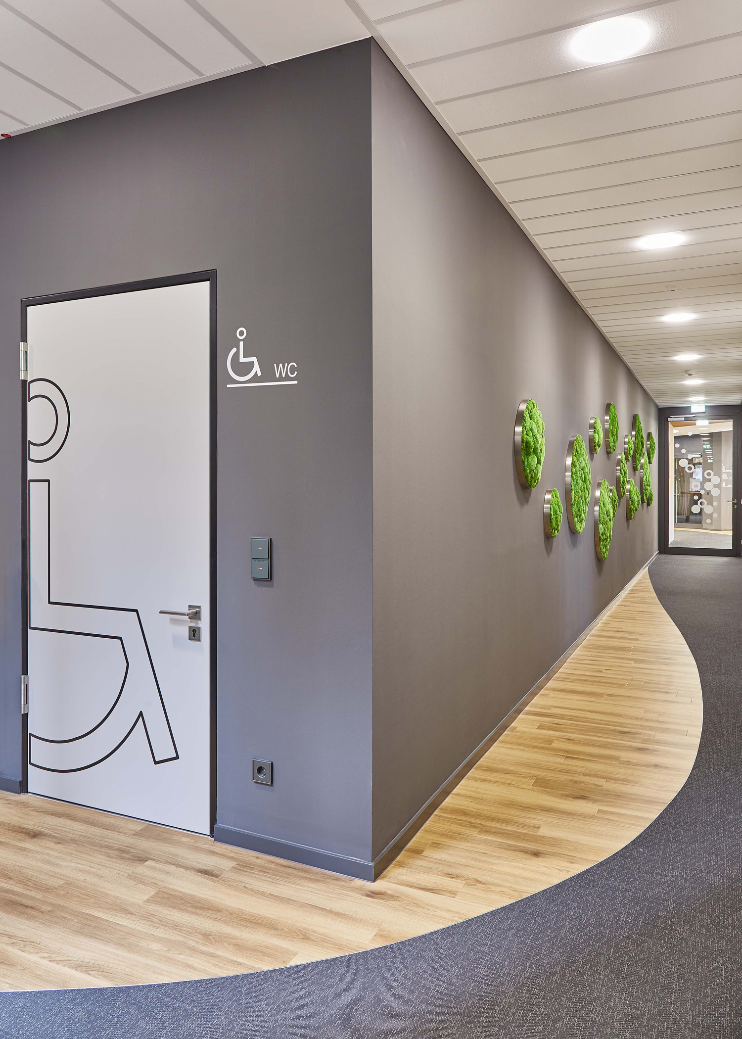 Foto: Moderner Flur mit grauer Wand, Parkett- und Teppichboden, runden Pflanzenwand Elemente, und WC Tür mit Signaletik Folierung.