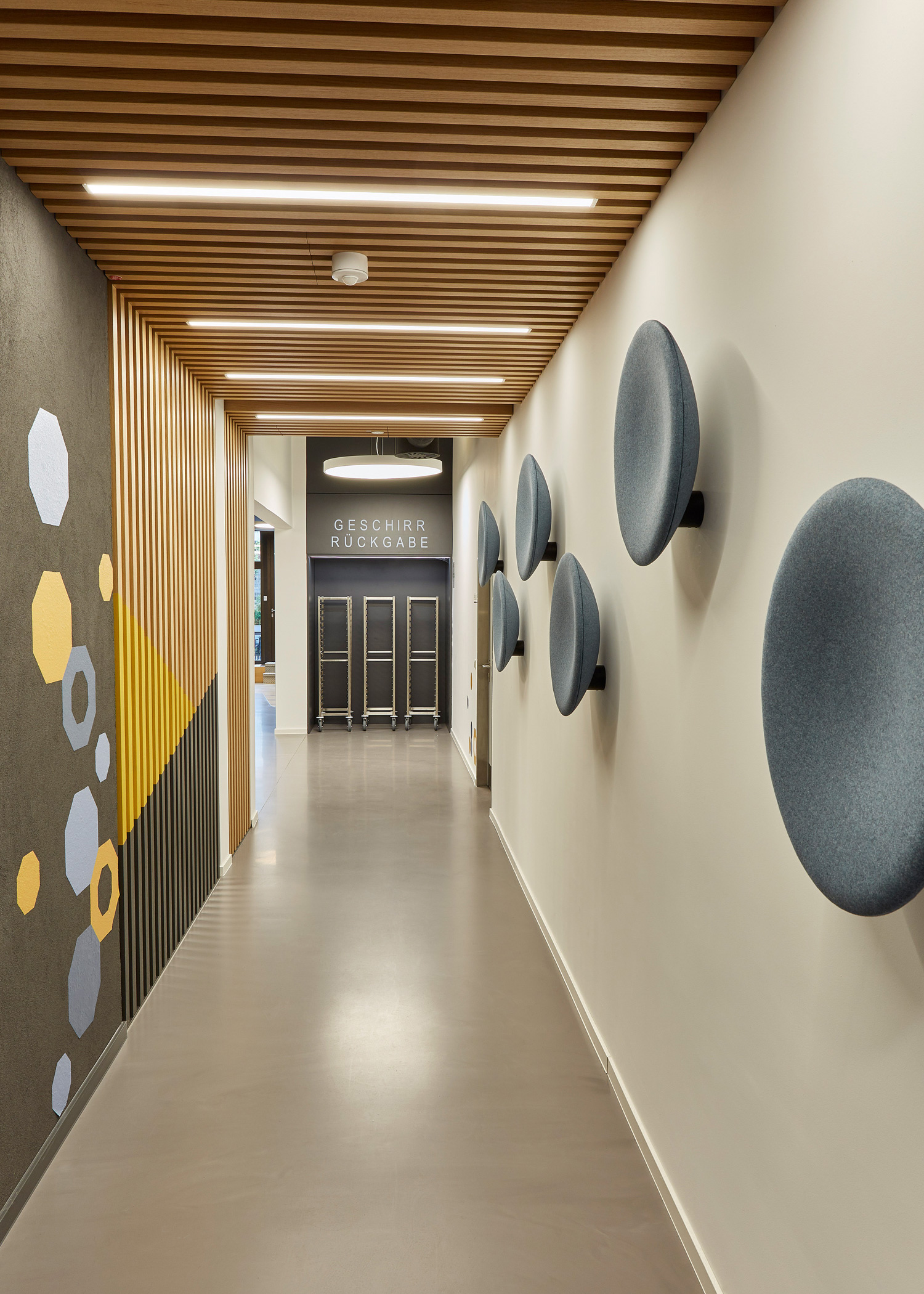 Foto: Modern gestalteter Flur mit Holzlamellen Wand- und Deckenverkleidung, Integrierter Beleuchtung, geometrischen Mustern in blau und Gelb, sowie blauen runden Akustikelementen an den Wänden.