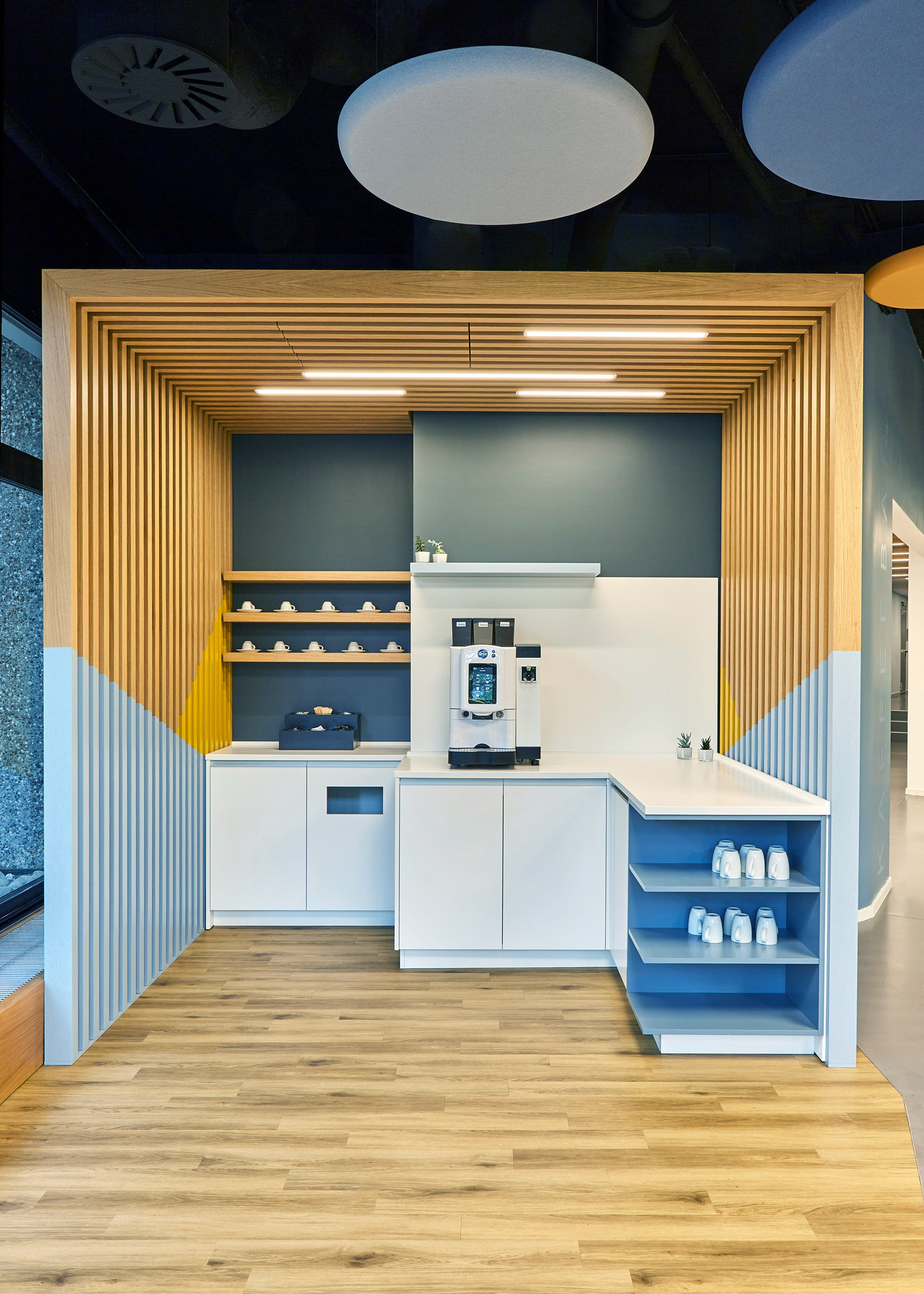 Foto: Modern gestaltete Kaffeestation mit heller Theke, Kaffeemaschine, offenen Regalen und einer markanten Holzlamellenkonstruktion. Blaue und gelbe Farbakzente sowie runde Akustikelemente an der Decke.