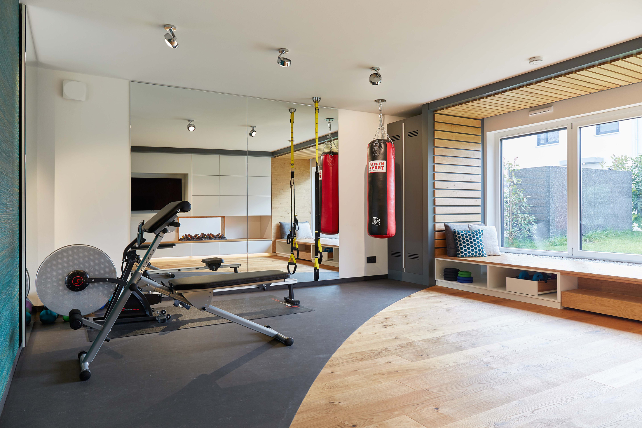 Foto: Home-Gym Fitnessbereich mit Sportgeräten, großer Fensterfront mit eingebauter Sitznische und Wandspiegel. Zonieren des Raumes durch verschiedene Bodenbeläge, warme Naturmaterialien.