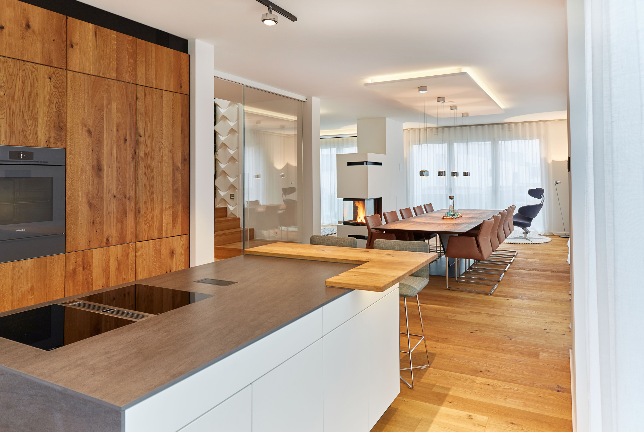 Foto: Heller, offen gestalteter Wohn- und Essbereich mit hochwertiger Holzküche, langem Esstisch, Kamin und bodentiefen Vorhängen in modernem Design.