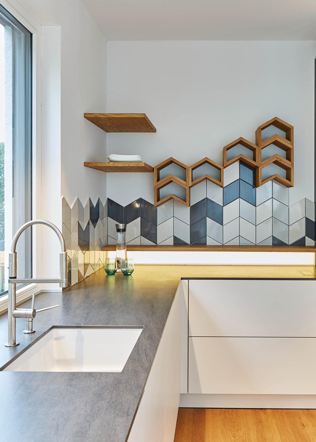 Foto: Moderne weiße Küche mit Arbeitsfläche in Anthrazit  und blau-weißen Fliesen in geometrischem Muster als Spritzschutz, Maßgefertigte Holzregale in gleicher Form mit indirekter Unterbeleuchtung.