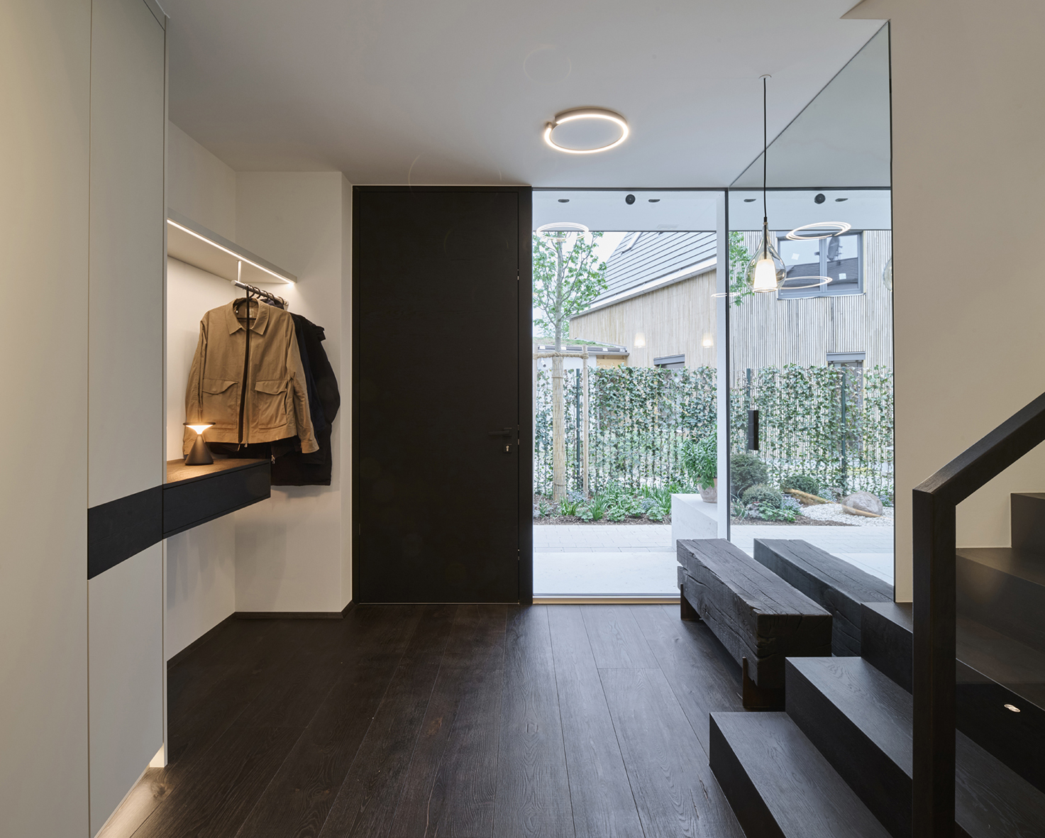 Foto: Moderner, lichtdurchfluteter Eingangsbereich mit Garderobe, integrierter Beleuchtung, dunkler Holztreppe, Spiegel und großen Fensterflächen.
