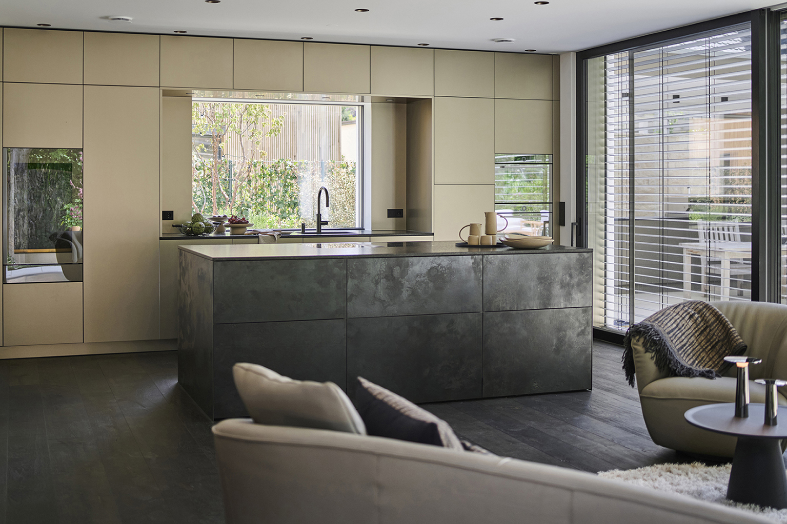Foto: Moderne, offen gestaltete Wohnküche mit zentraler Kochinsel in dunkler Metalloptik, deckenhohen, cremefarbenen Schrankfronten und großflächigen Fensterfronten. Warme Naturtöne, dunkler Holzboden.
