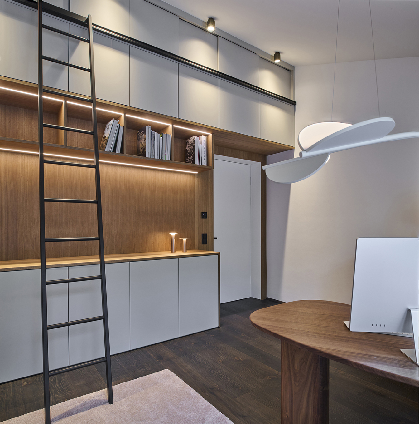 Foto: Modernes, hochwertiges, maßgefertigtes Homeoffice mit deckenhohen Einbauschränken in grau und Holzoptik, integrierter LED-Beleuchtung, schwarzer Leiter, dunklem Holzboden und organisch geformtem Schreibtisch.