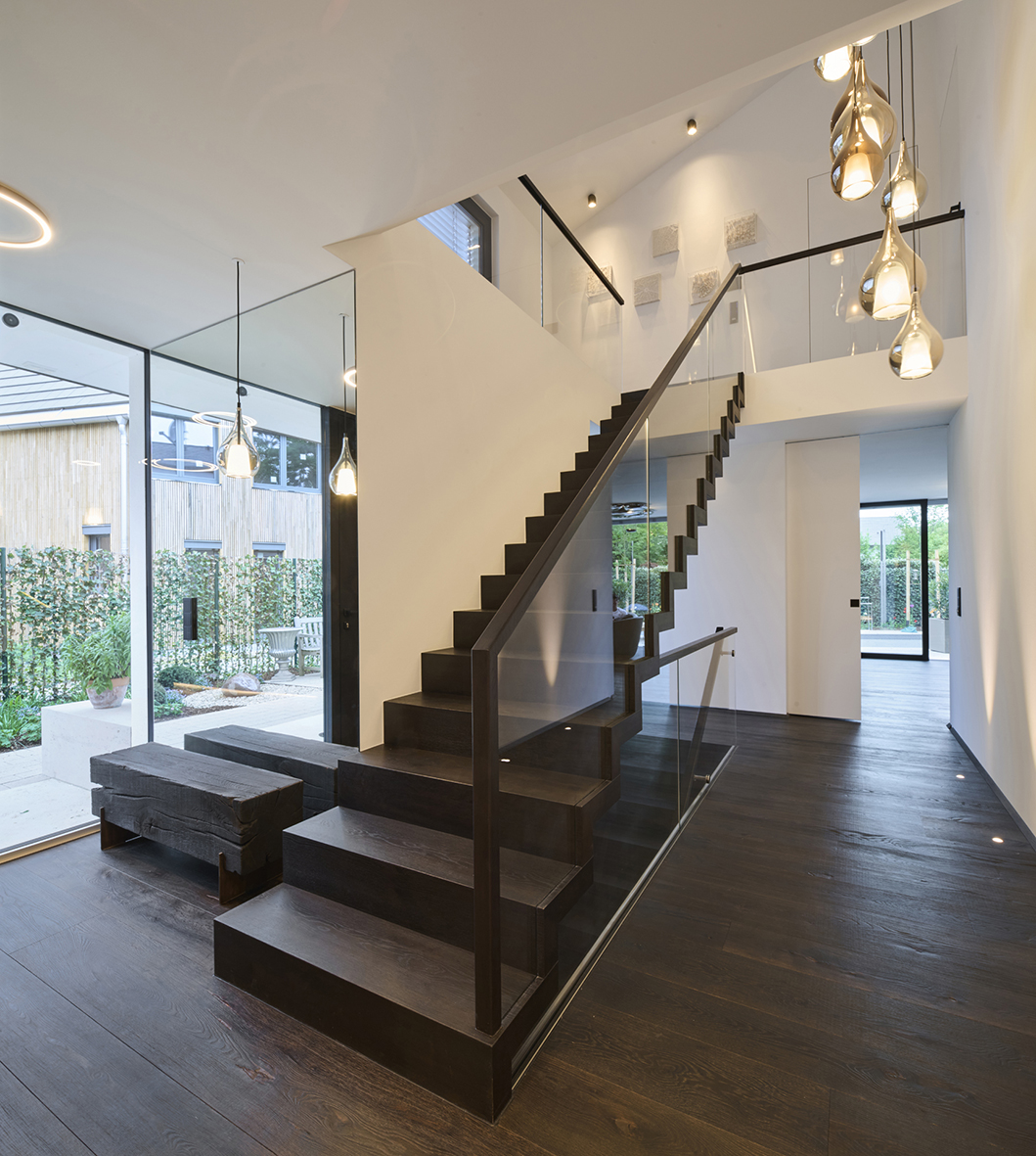 Foto: Moderner, lichtdurchfluteter Eingangsbereich mit freitragender Holztreppe, Glasgeländer, dunklem Holzboden, tropfenförmigen Pendelleuchten und großen Fensterflächen.