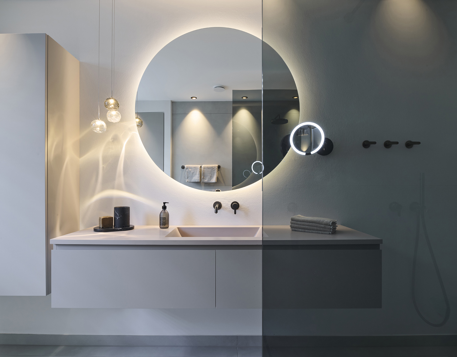 Foto: Modernes, hochwertig gestaltetes Badezimmer mit freischwebendem Waschtisch, großem rundem Spiegel und eleganten Glaspendelleuchten, deren Lichtreflexionen eine warme und luxuriöse Atmosphäre schaffen.