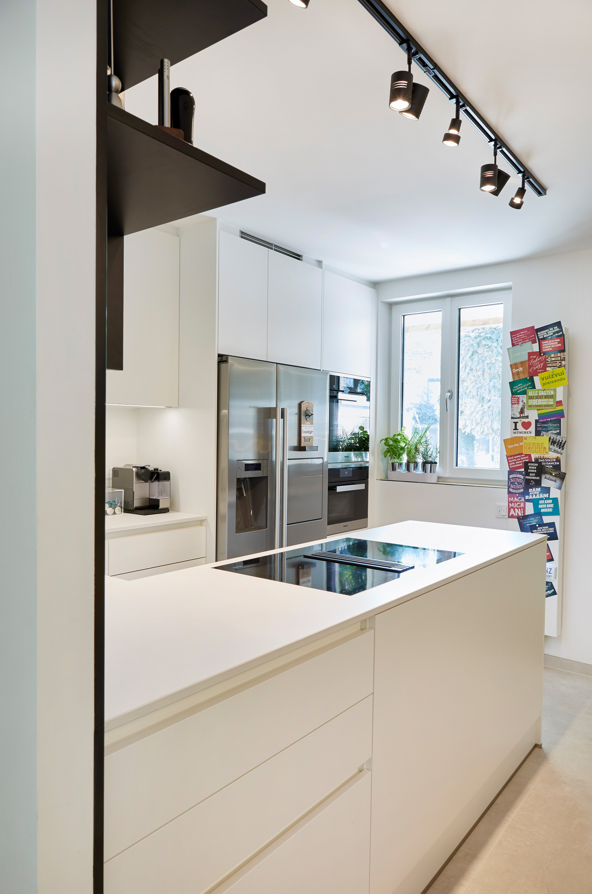 Foto: Minimalistische, helle, weiße Küche mit in Einbauschränken integrierten Küchengeräten, Pinnwand mit Postkarten und Beleuchtung durch Spots an Deckenschiene. 