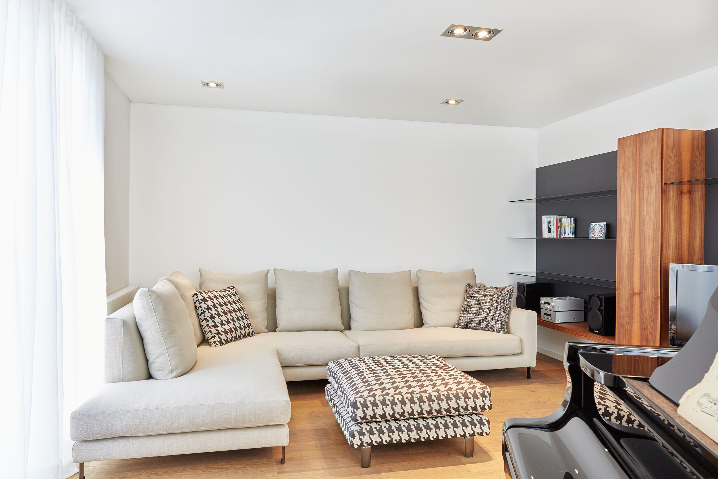 Foto: Heller, modern eingerichteter Wohnraum mit großem cremefarbenem Ecksofa, gemustertem Hocker, Holzboden und minimalistischen Regalen neben einem schwarzen Klavier.