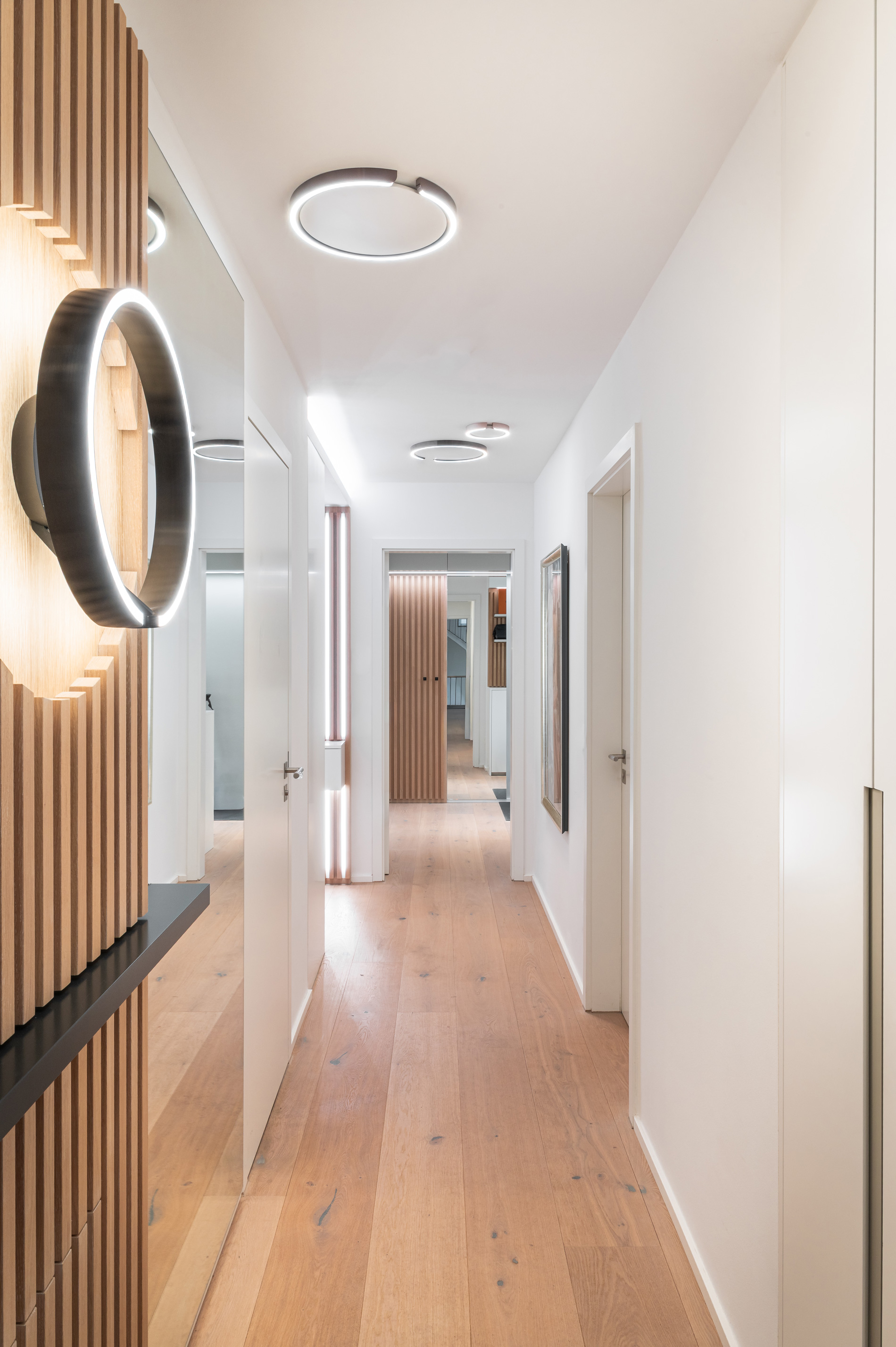 Foto: Heller, minimalistischer Flur mit hellem Holzboden, weißen Wänden, deckenmontierten Ring-Leuchten und vertikalen Holzlamellen mit integriertem Spiegel.